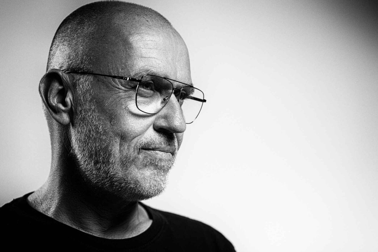 Black and white portrait of a bald man with a beard and glasses, wearing a black shirt, looking thoughtfully to the side.